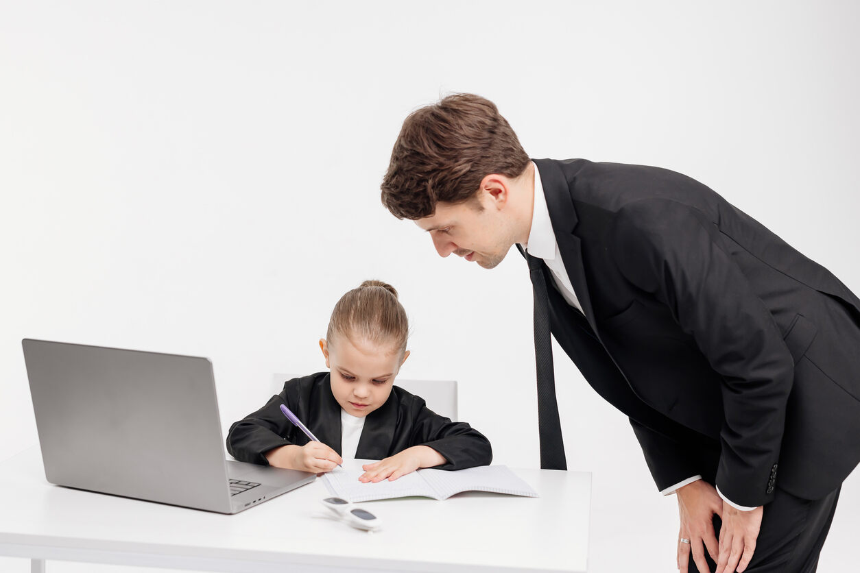 A man in a suit attentively watches a young girl writing at a table with a laptop, symbolizing the role of a Guardian Ad Litem lawyer observing a child in a Kansas child custody case.
